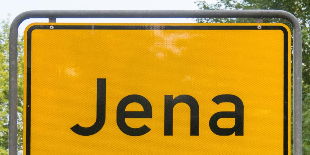 Ein gelbes Ortseingangsschild mit dem Schriftzug Jena vor einem Naturhintergrund