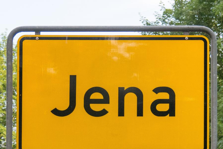 Ein gelbes Ortseingangsschild mit dem Schriftzug Jena vor einem Naturhintergrund